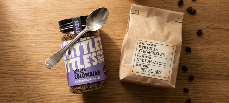 A split flat-lay image: on the left, a premium instant coffee jar (like Little's) with a spoon resting on it; on the right, a small kraft bag of single-origin specialty coffee beans with visible roast date. Clean wooden surface, natural light.